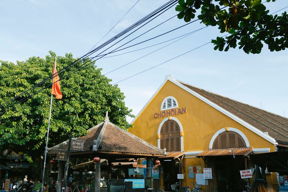A traditional yellow market building in Hoi An, Vietnam, with lush greenery and a Vietnamese flag.