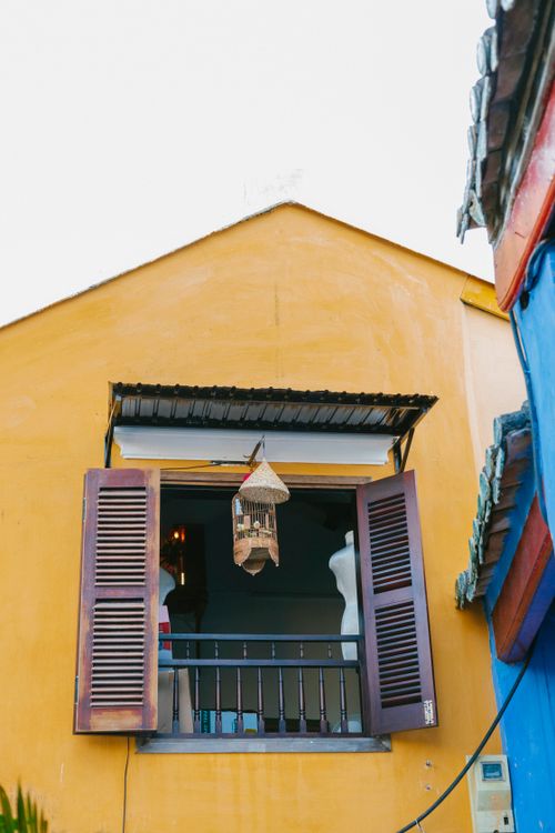 Free Yellow house facade with an open window and a hanging birdcage, creating a quaint street scene. Stock Photo