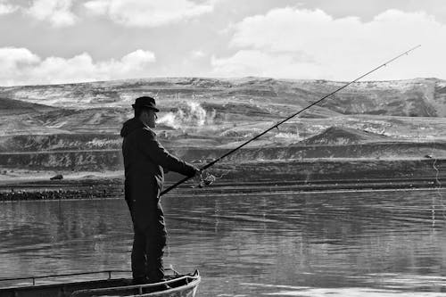 Grayscale Photo of Man in Black Jacket Standing on Boat and Fishing 