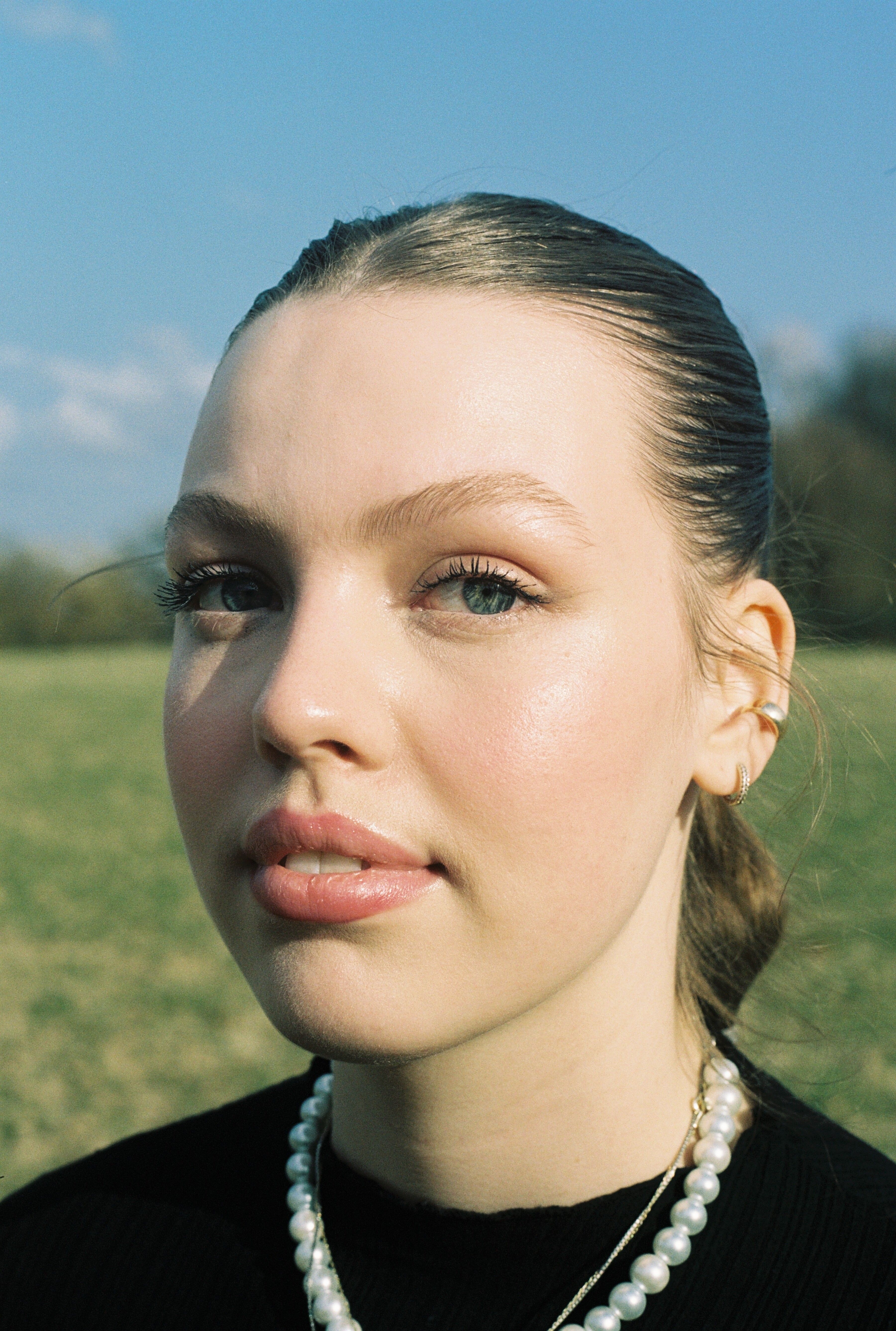 Close-up portrait highlighting a woman's natural beauty with a pearl necklace against a serene outdoor backdrop.