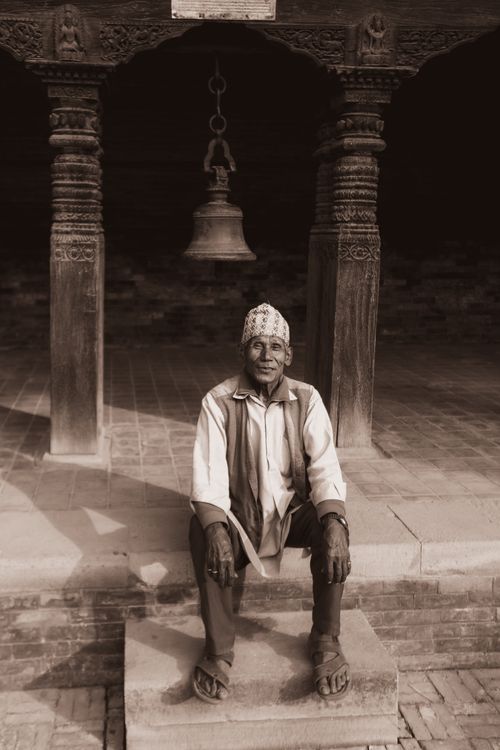 Free An elderly man sits contently at the entrance of a historical temple, captured in sepia tones. Stock Photo