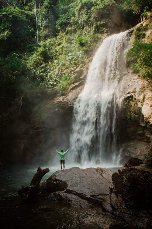 Free Person Standing on a Rock by a Waterfall with His Arms Raised  Stock Photo