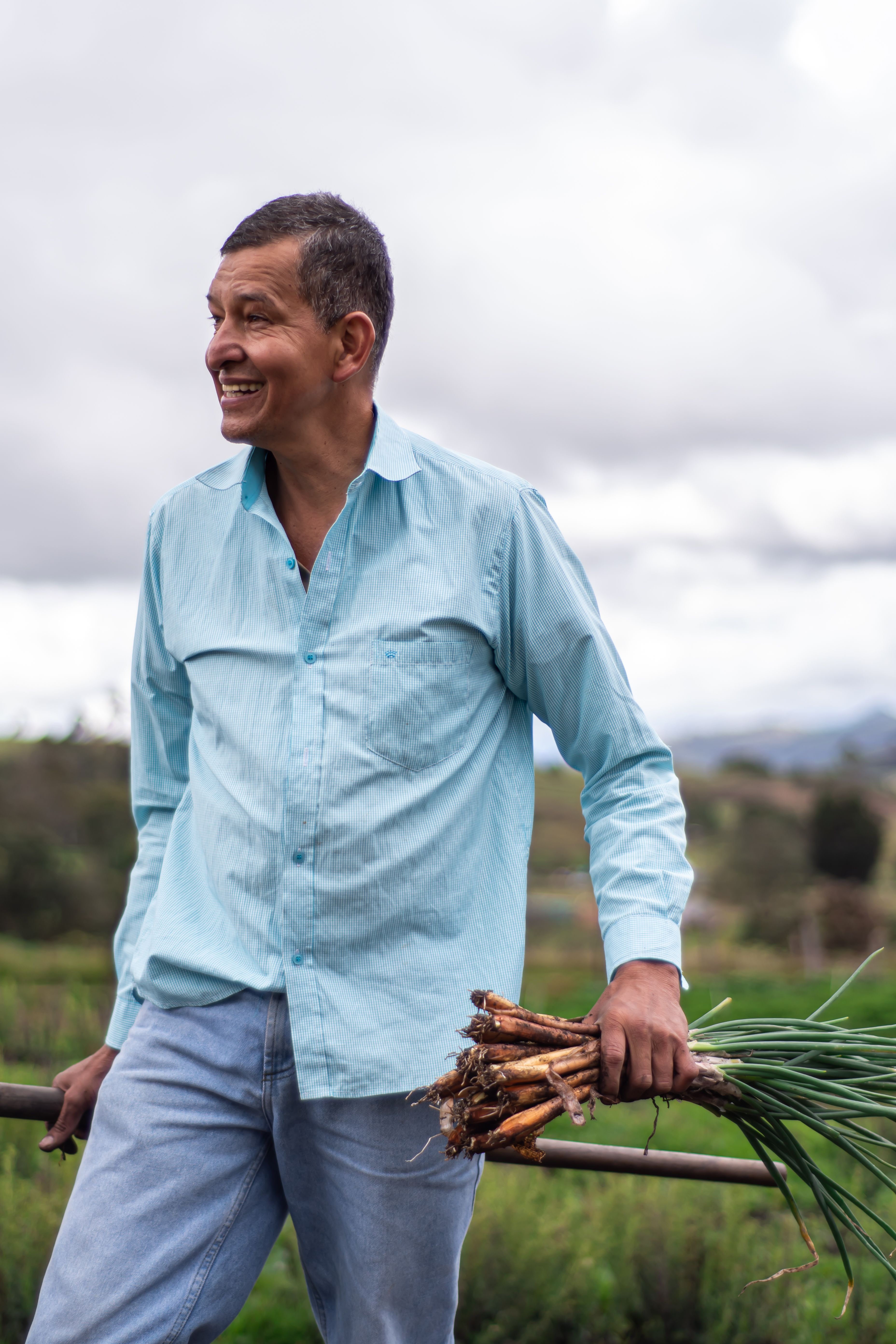 A farmer in a blue shirt holds fresh scallions, smiling in a countryside field.