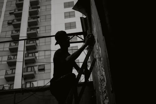 Grayscale Photo of Man With Cap Standing on Ladder