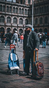 Senior man wearing a mask and standing with trolley bags in a bustling Bogotá marketplace.