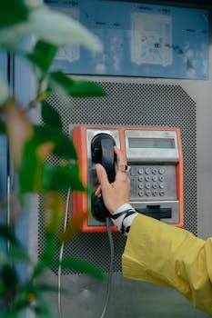 Close-up shot of a hand holding the receiver of a vintage orange payphone amid greenery.