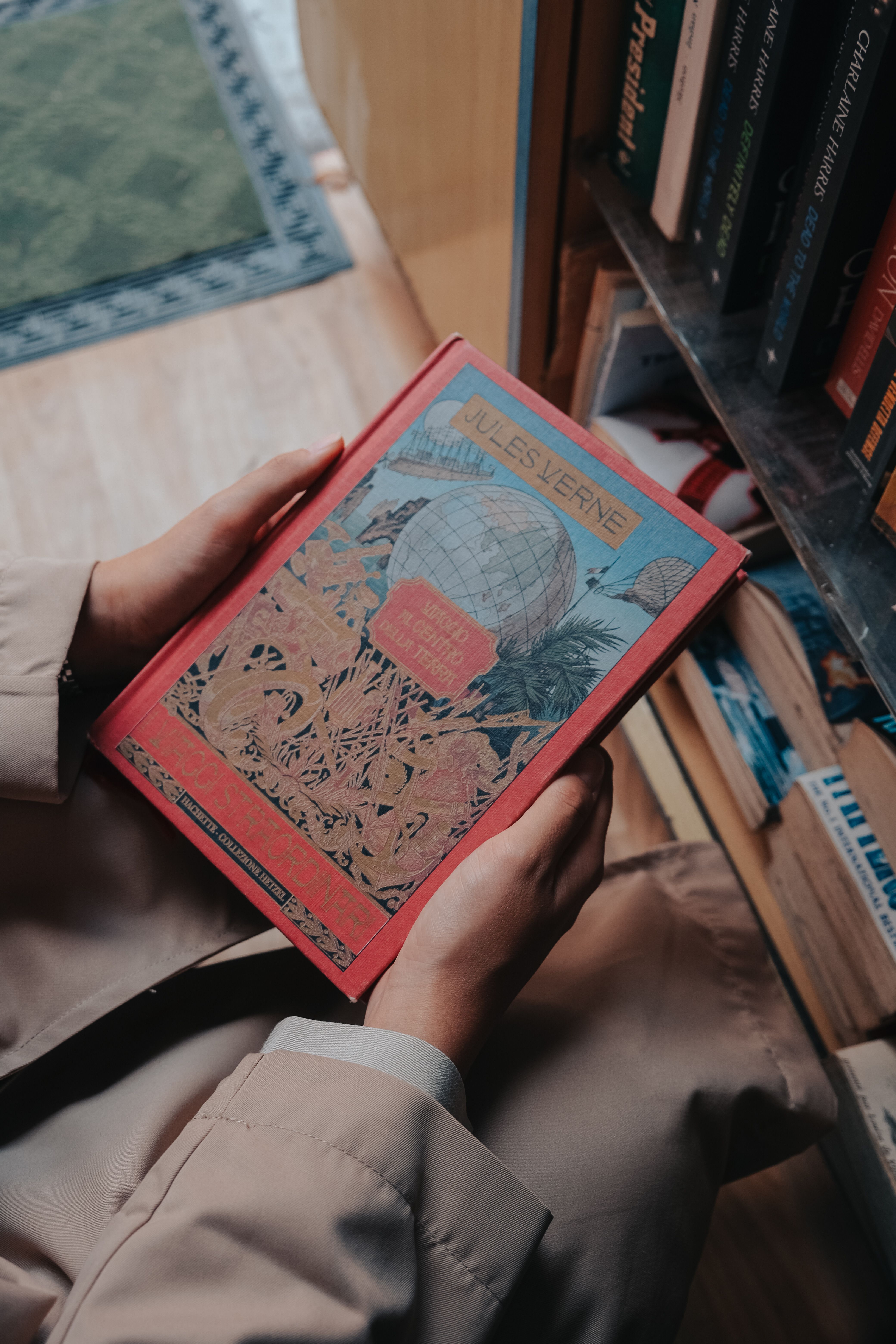 A close-up view of someone holding a colorful Jules Verne book in a cozy library.