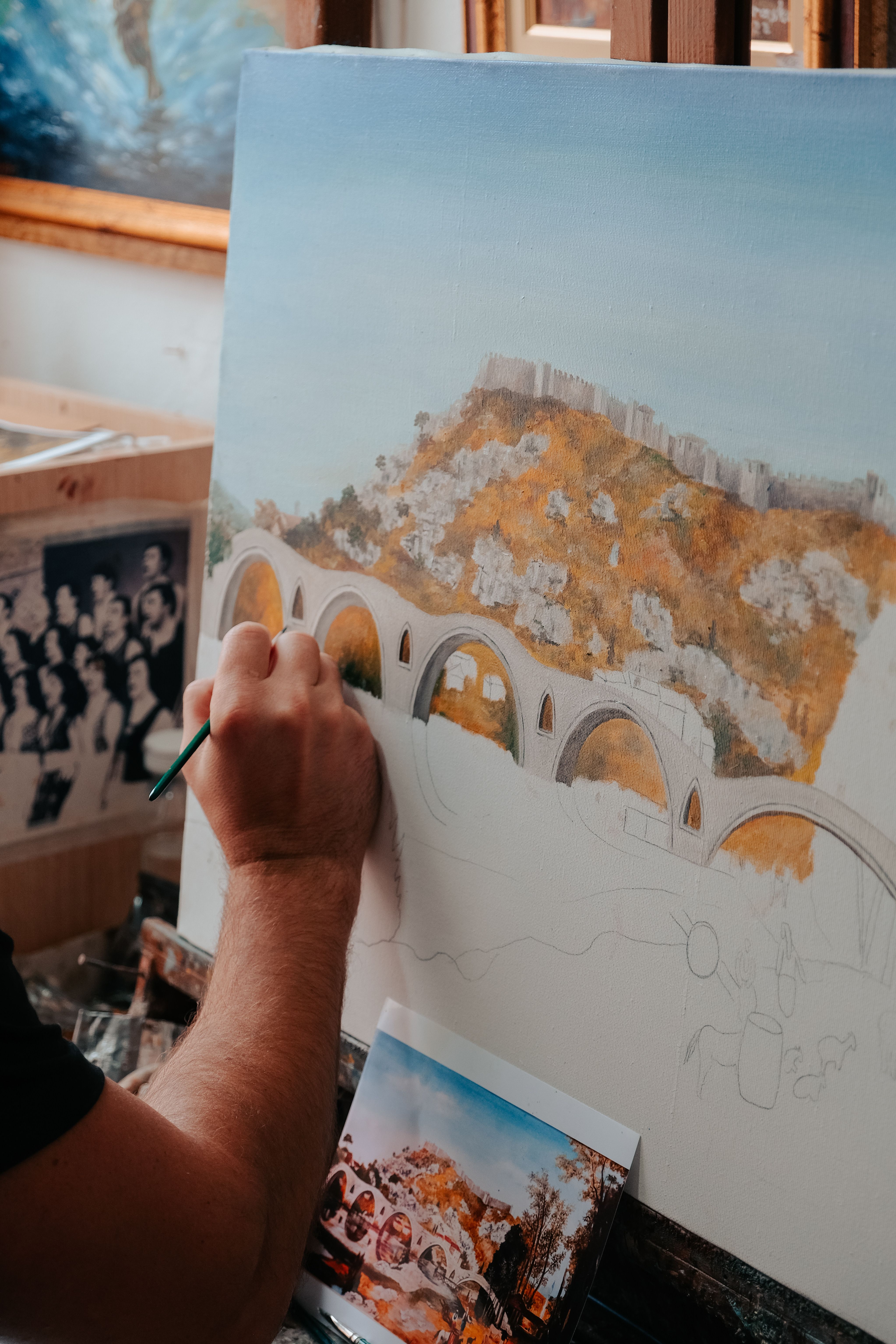 Artist hand painting a scenic landscape featuring an arched bridge and hill with brush on canvas.