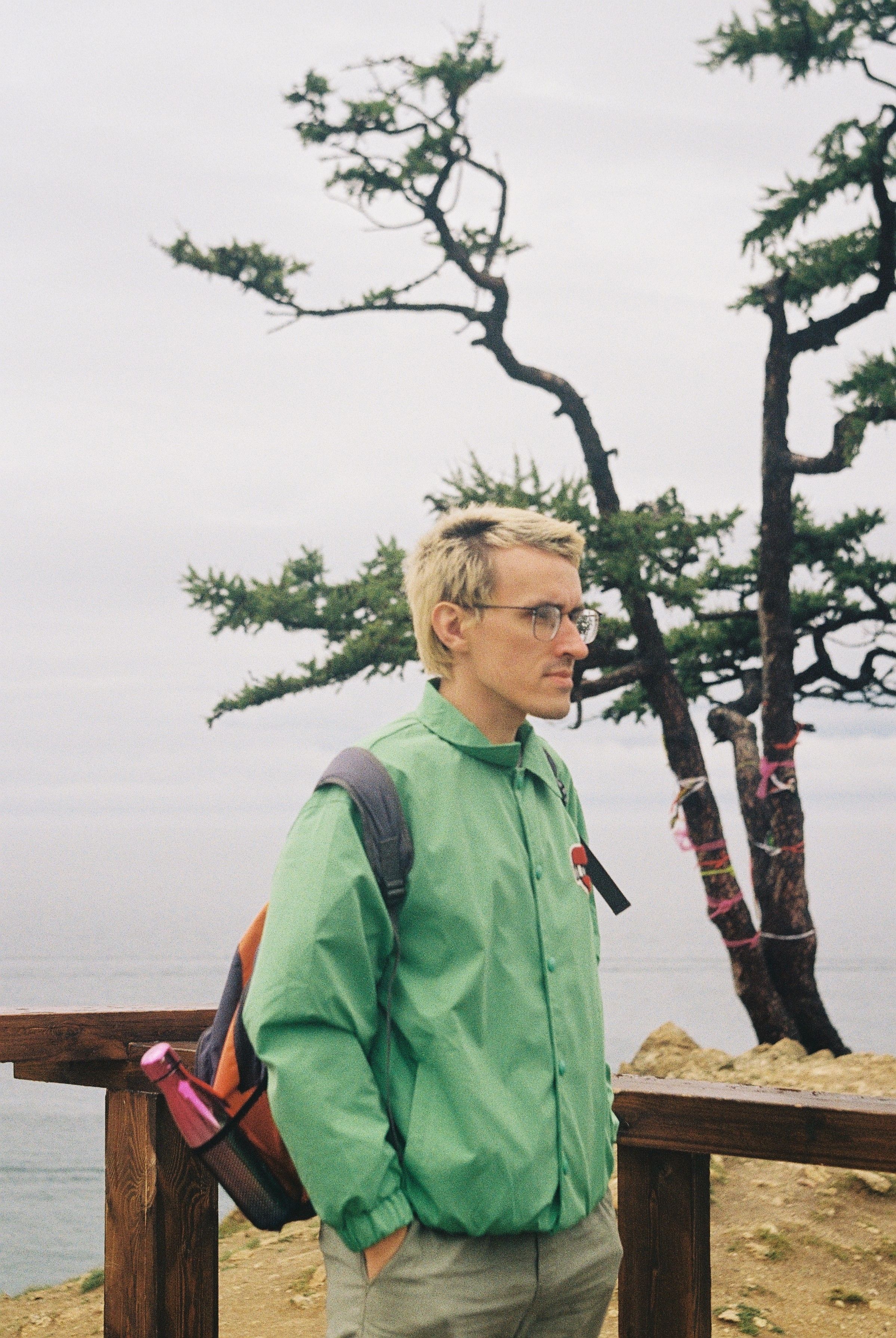 A person wearing a green jacket stands by a coastal area with a backpack and a tree in the background.