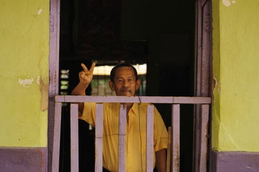 Elderly man in yellow shirt posing with peace sign at doorway in Carmen de Apicalá, Colombia.