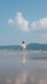 A woman walks leisurely on a serene beach with mountains in the background and a clear blue sky overhead.