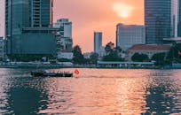 日落时分，一艘小船沿着城市海滨行驶，城市的摩天大楼倒映在平静的河面上