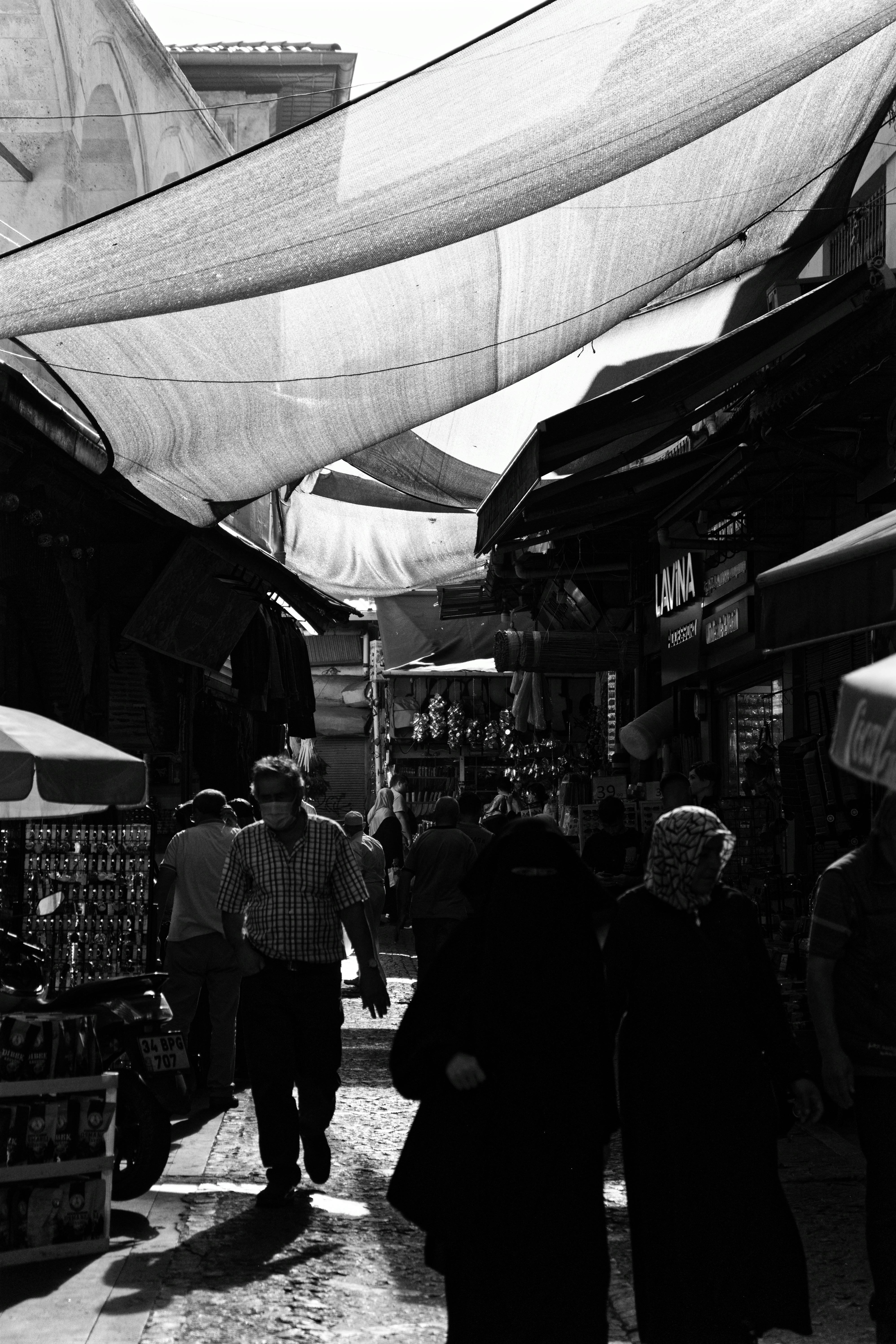 Black and white photo of a bustling market street with people walking under fabric awnings.
