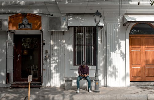 A Man in Red Long Sleeves and Black Cap Sitting on a Concrete Bench Near a Street Light and White Building While Using a Phone