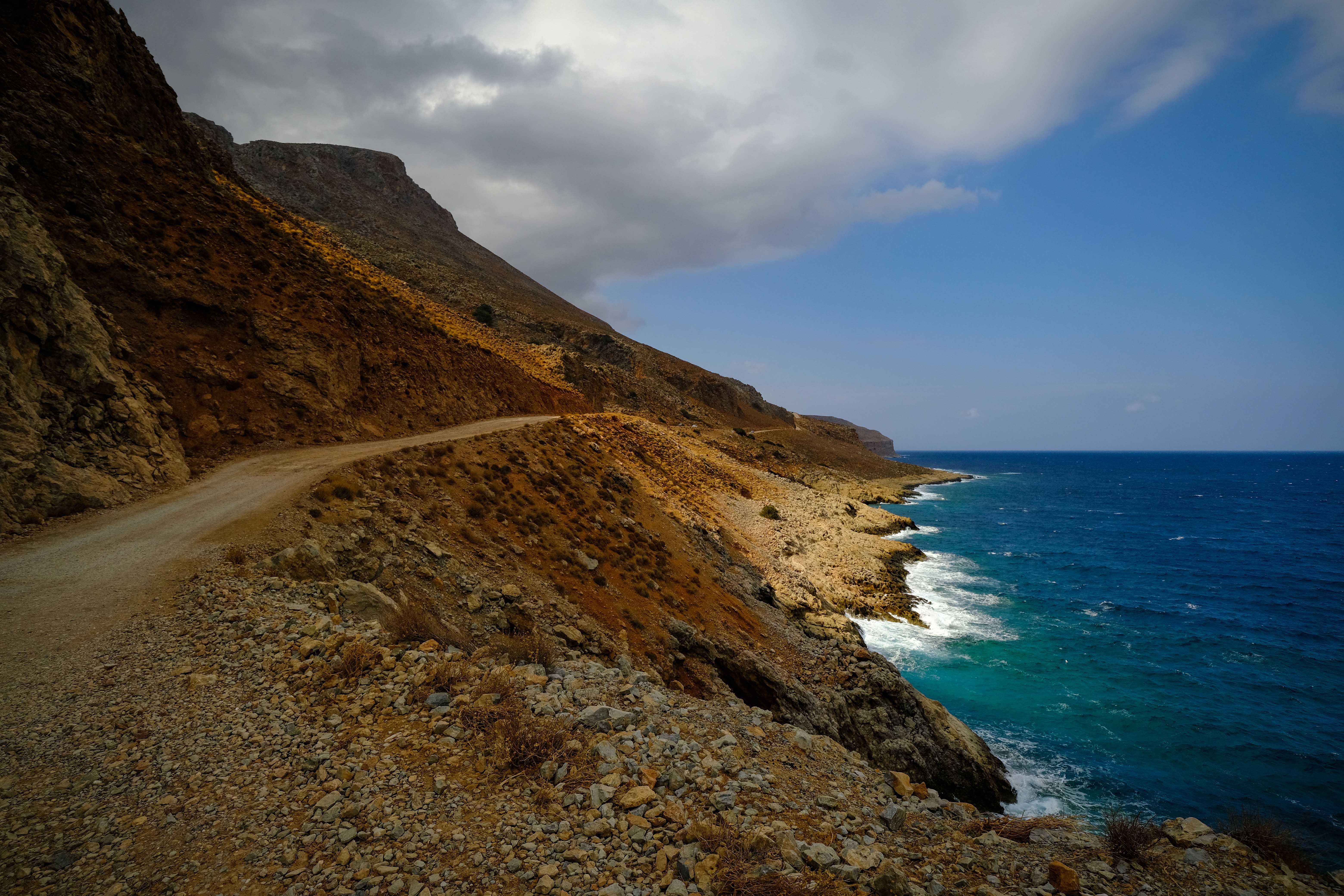 Rustic coastal road winding along a rocky cliff with ocean views under a blue sky. - Photo by Francesco Ungaro on Pexels