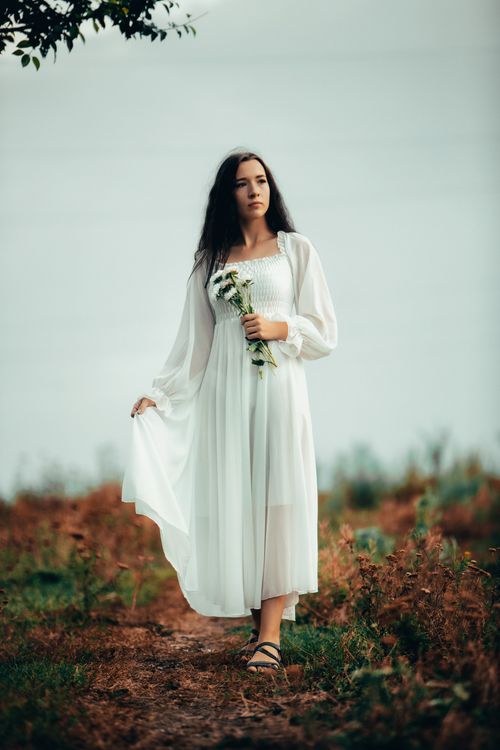 Free A young woman in a white dress holding flowers, standing in a natural outdoor setting. Stock Photo