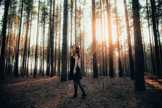 A woman in a dark dress stands in a sunlit forest in Ankara, Türkiye, surrounded by tall trees.