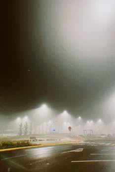 A foggy night captures illuminated streetlights casting a moody atmosphere on an urban road.