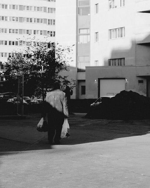 Grayscale Photography of Man Walking on Street Holding Plastic Bags