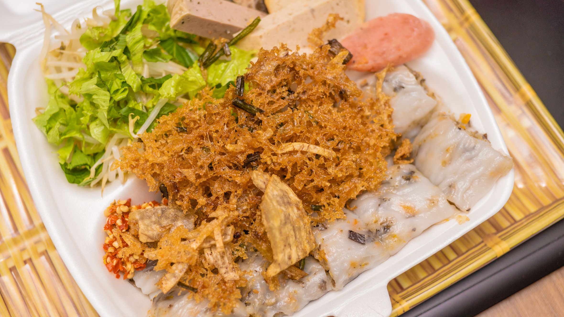 A tempting close-up of traditional Vietnamese bánh cuốn in a takeaway box with fresh greens and sausage.