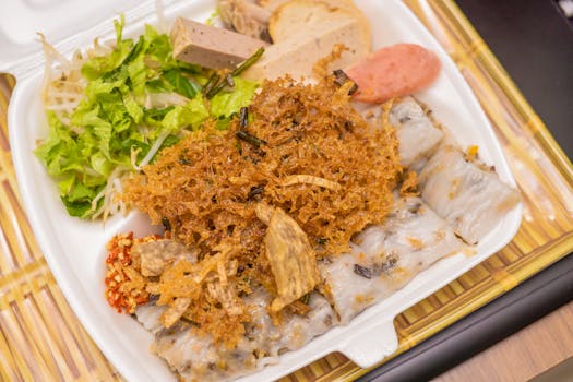 A tempting close-up of traditional Vietnamese bánh cuốn in a takeaway box with fresh greens and sausage.