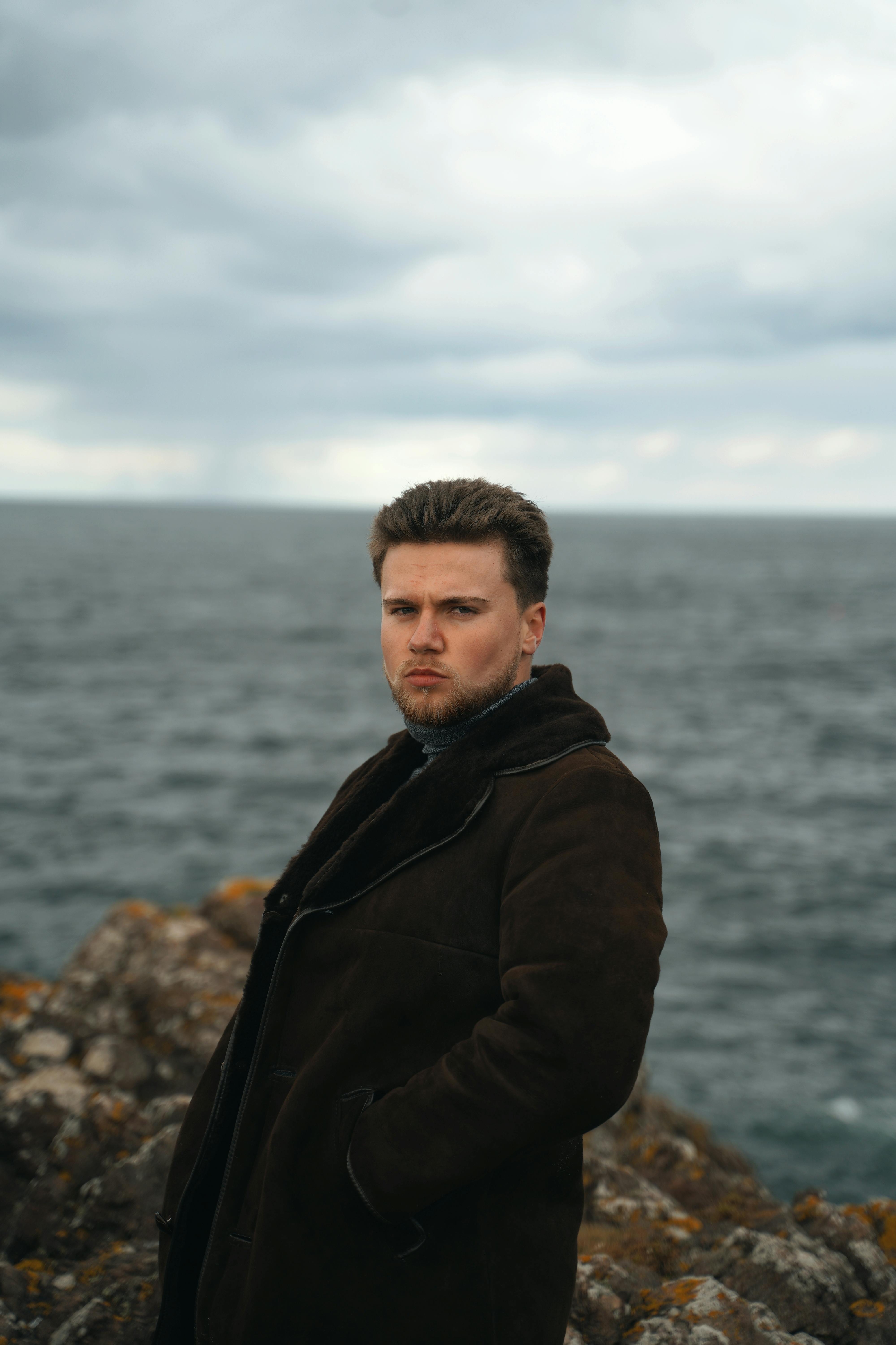 Portrait of a serious man in a brown jacket standing by the ocean.