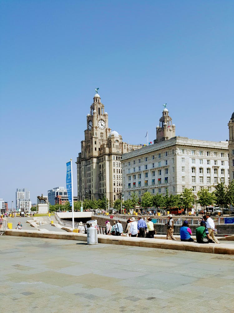 Group Sitting On A Sidewalk Looking At The Royal Liver Building In Liverpool, England