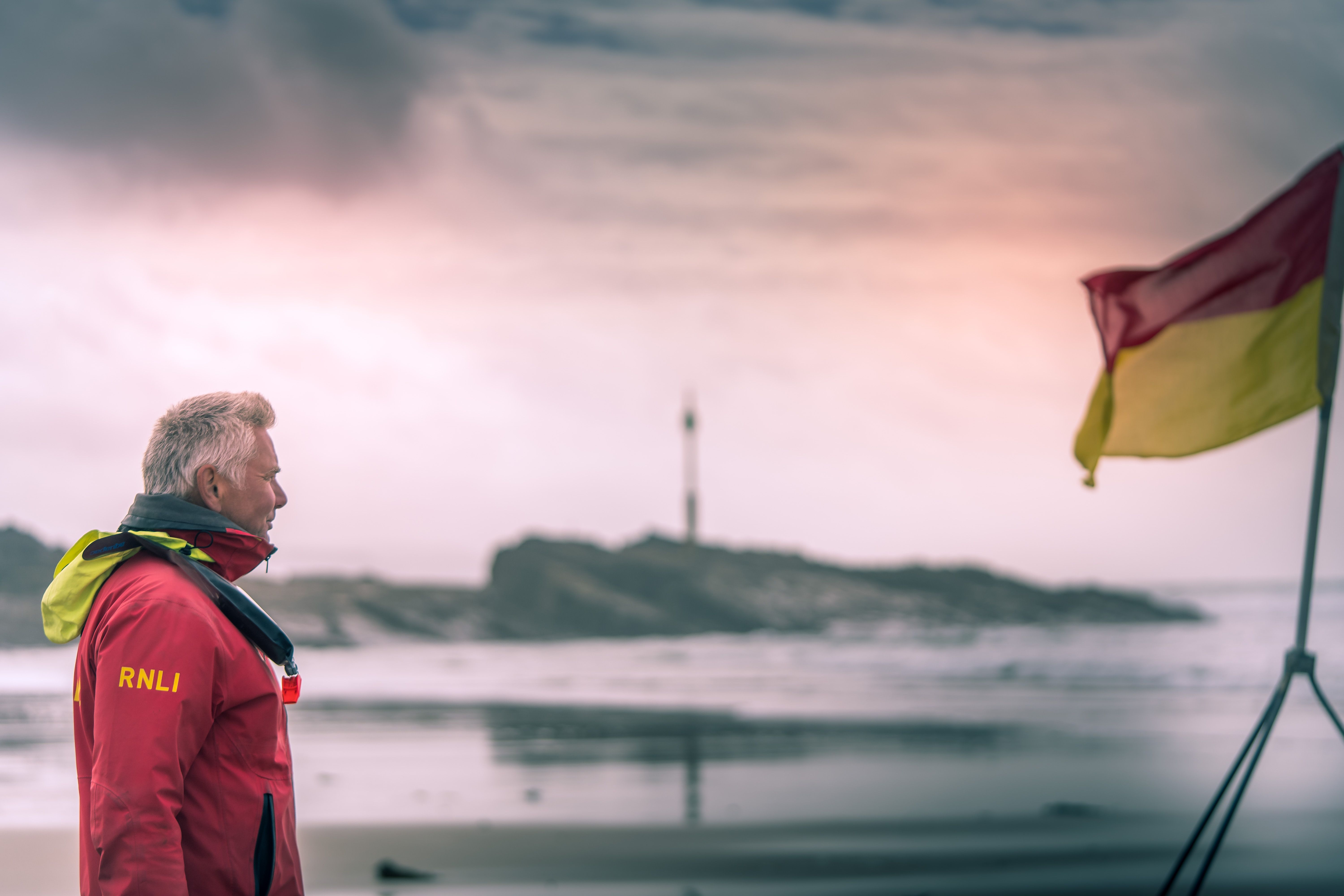 Lifeguard standing by the ocean shore during sunrise, wearing a red RNLI jacket, flag billowing in the breeze.