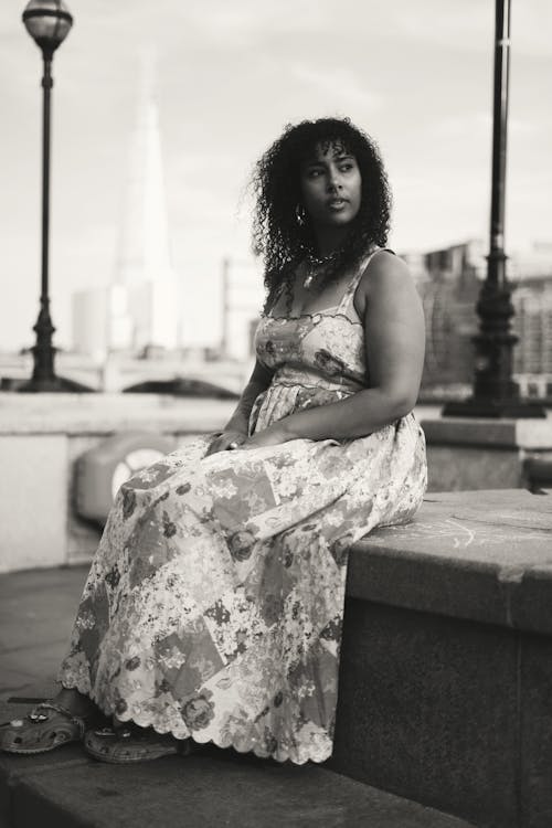 A Grayscale Photo of a Woman in Printed Dress Sitting on a Concrete Bench