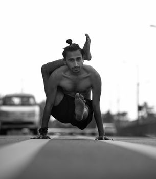 A shirtless man in a monochrome image demonstrating an advanced yoga pose on the road.