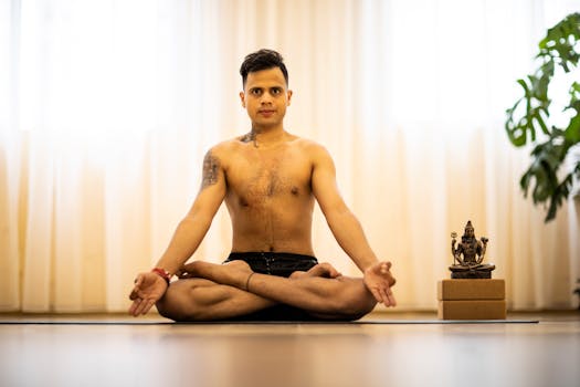 Man meditating in a lotus pose indoors with a statue beside him, promoting mindfulness.