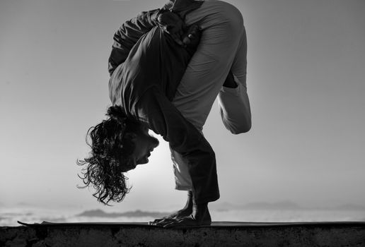 A black and white photo of a person demonstrating extreme flexibility in a yoga pose outdoors.