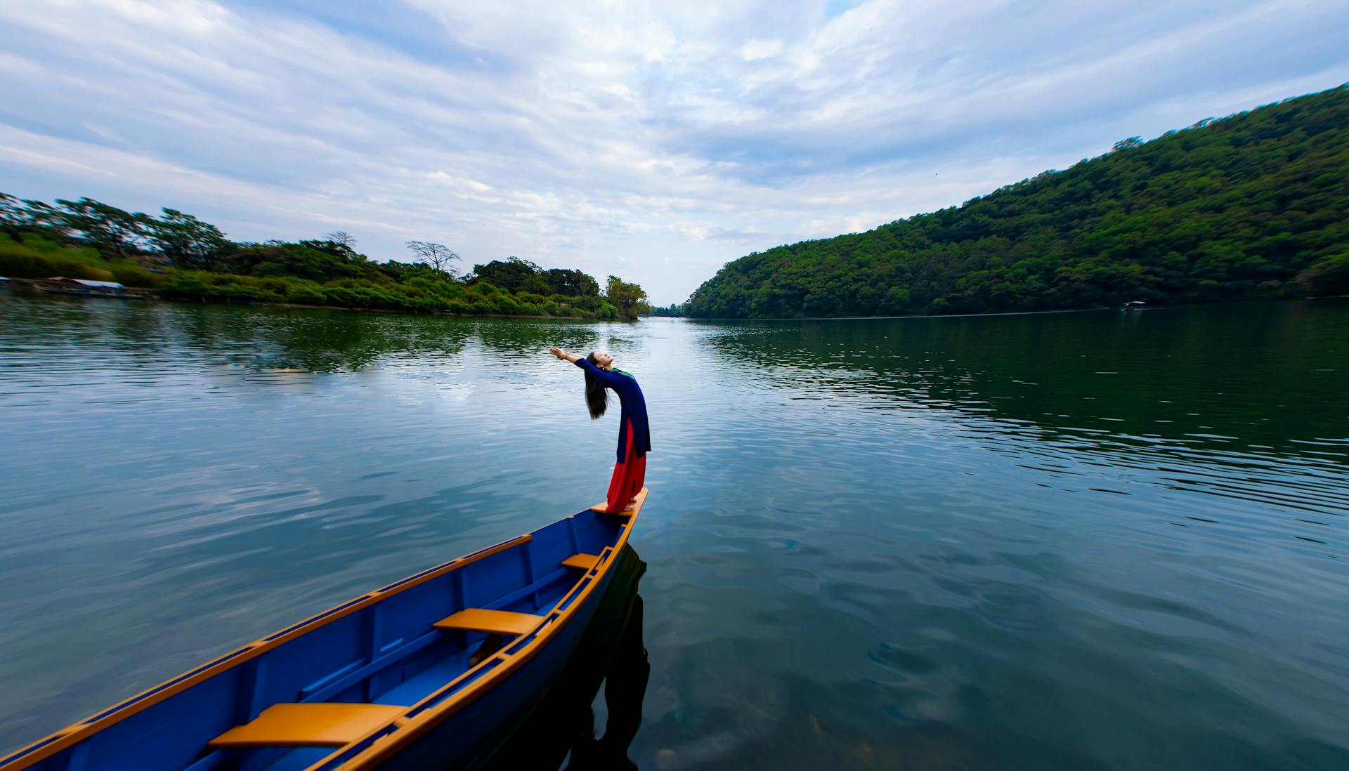 A person gracefully balancing on a boat surrounded by a peaceful river and lush green hills under a cloudy sky.