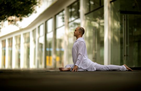 A man in white clothing performs a yoga stretch outdoors, showcasing relaxation and focus.