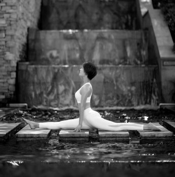 Woman performing a split yoga pose near a serene waterfall in monochrome.