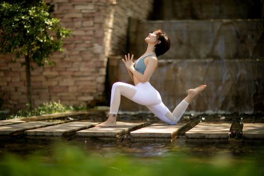 Woman performing yoga pose by a serene waterfall and stone path outdoors.
