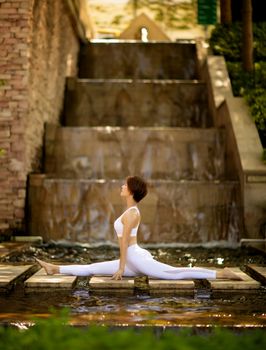 Woman performing yoga split on stepping stones by a cascading waterfall outdoors.