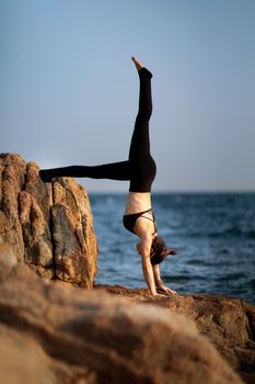 A woman in activewear performing a handstand on seaside rocks, showcasing balance and strength.