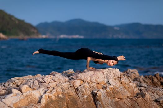 Asian woman performs yoga pose on rocky seashore with mountainous backdrop.