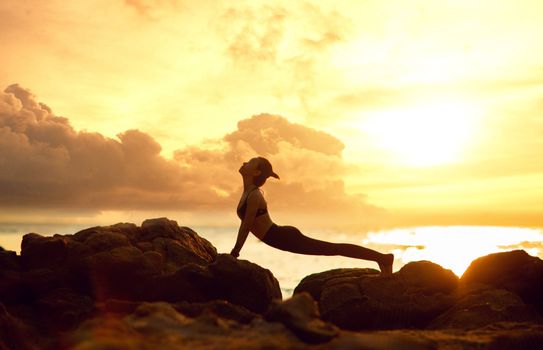 Silhouette of a woman doing yoga on a rocky beach at sunset, perfect for relaxation themes.