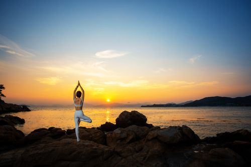 Free A woman performing yoga on rocky coastline at sunset, promoting relaxation and wellbeing. Stock Photo