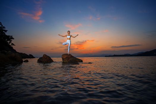 A woman performs yoga on rocks by a tranquil lake during a vibrant sunset.