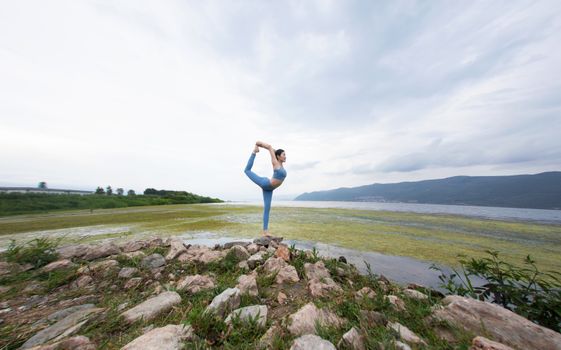 A woman practicing yoga on rocks by a serene lake with mountain backdrop.