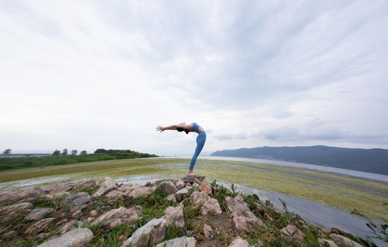 A woman in yoga attire stretches on rocks near a marshy landscape, under a cloudy sky.