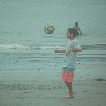 Child playing soccer on a sandy beach near the sea on a cloudy day.