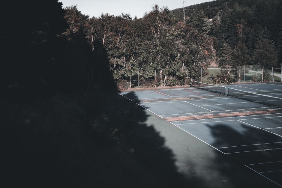 Empty outdoor tennis courts surrounded by trees, casting long shadows.