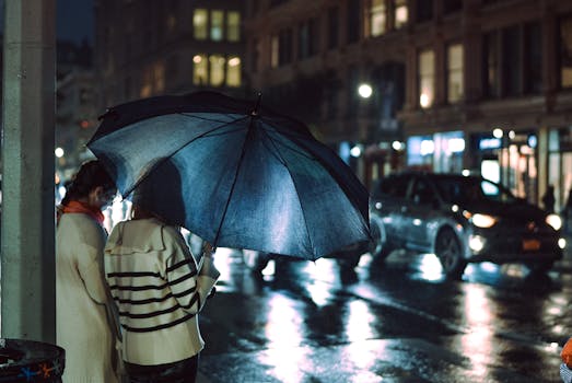 Women with umbrella on rainy city street at night, reflecting lights.