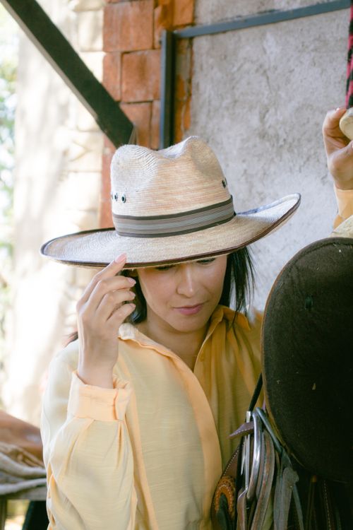 Free A woman adjusts her wide brim straw hat in a rustic barn setting, blending natural light and shadows. Stock Photo