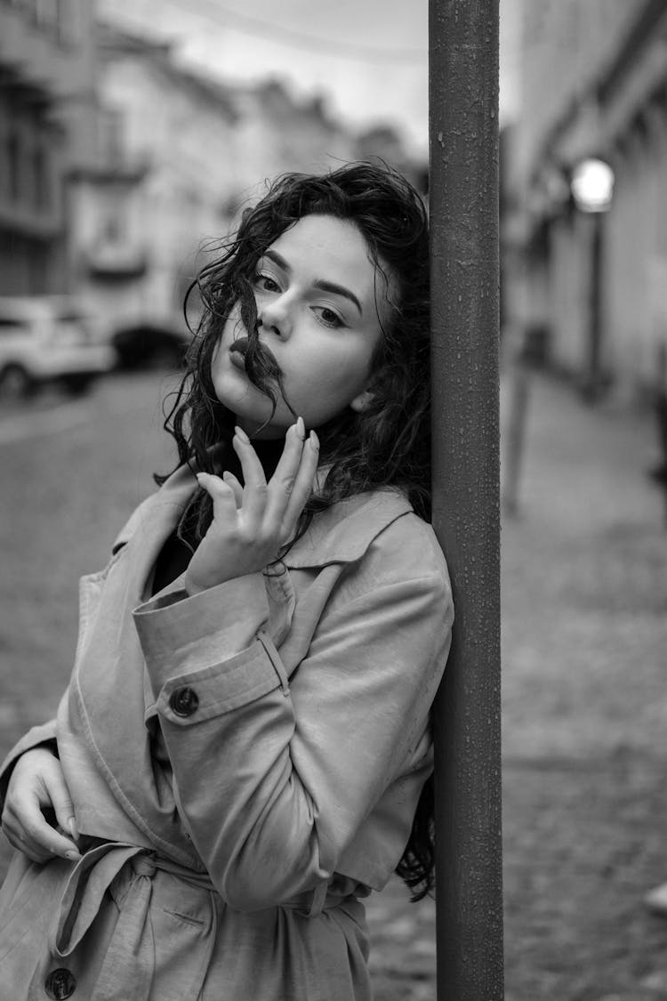 Grayscale Photo Of Woman In Coat Standing Beside A Pole 