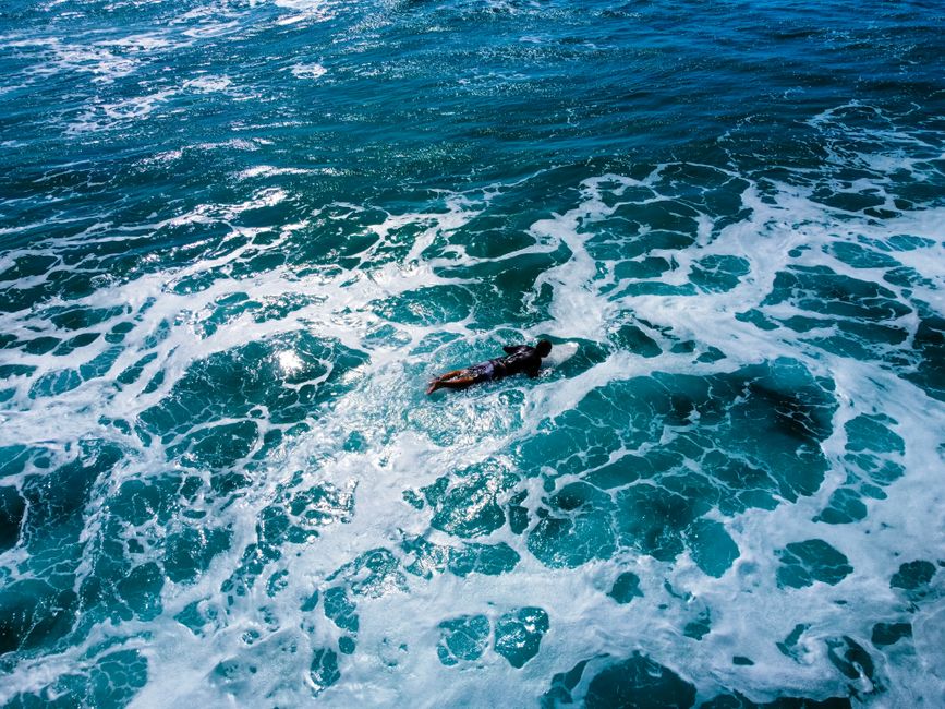 Aerial view of a surfer paddling through blue ocean waves with foamy textures.